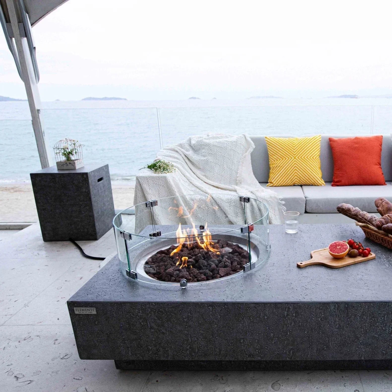 Elementi Metropolis fire pit table with glass windscreen on a patio by the ocean, modern outdoor seating