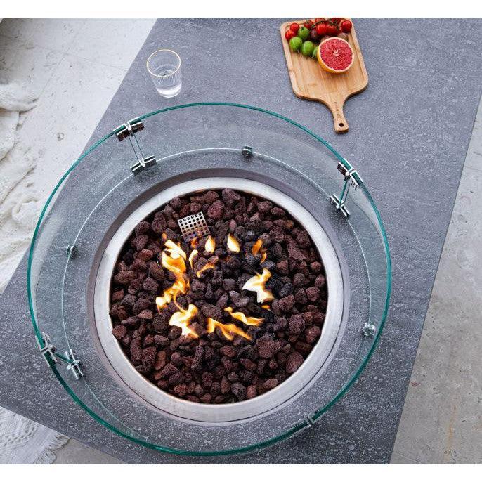Metropolis round outdoor fire pit table with glass wind guard, lava rocks, and visible flames