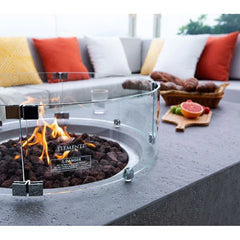 Elementi round outdoor gas fire pit with glass wind guard on patio table, modern seating and cushions